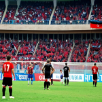 A vibrant football (soccer) match in Tirana, Albania. The stadium is packed with cheering fans waving red and black Albanian flags. In the foreground, a young boy wearing a Dinamo Tirana jersey gazes in awe at the players on the field. The scene captures the passion and excitement surrounding Albanian football, with a focus on the dreams of young athletes. Style: Realistic, dynamic lighting.